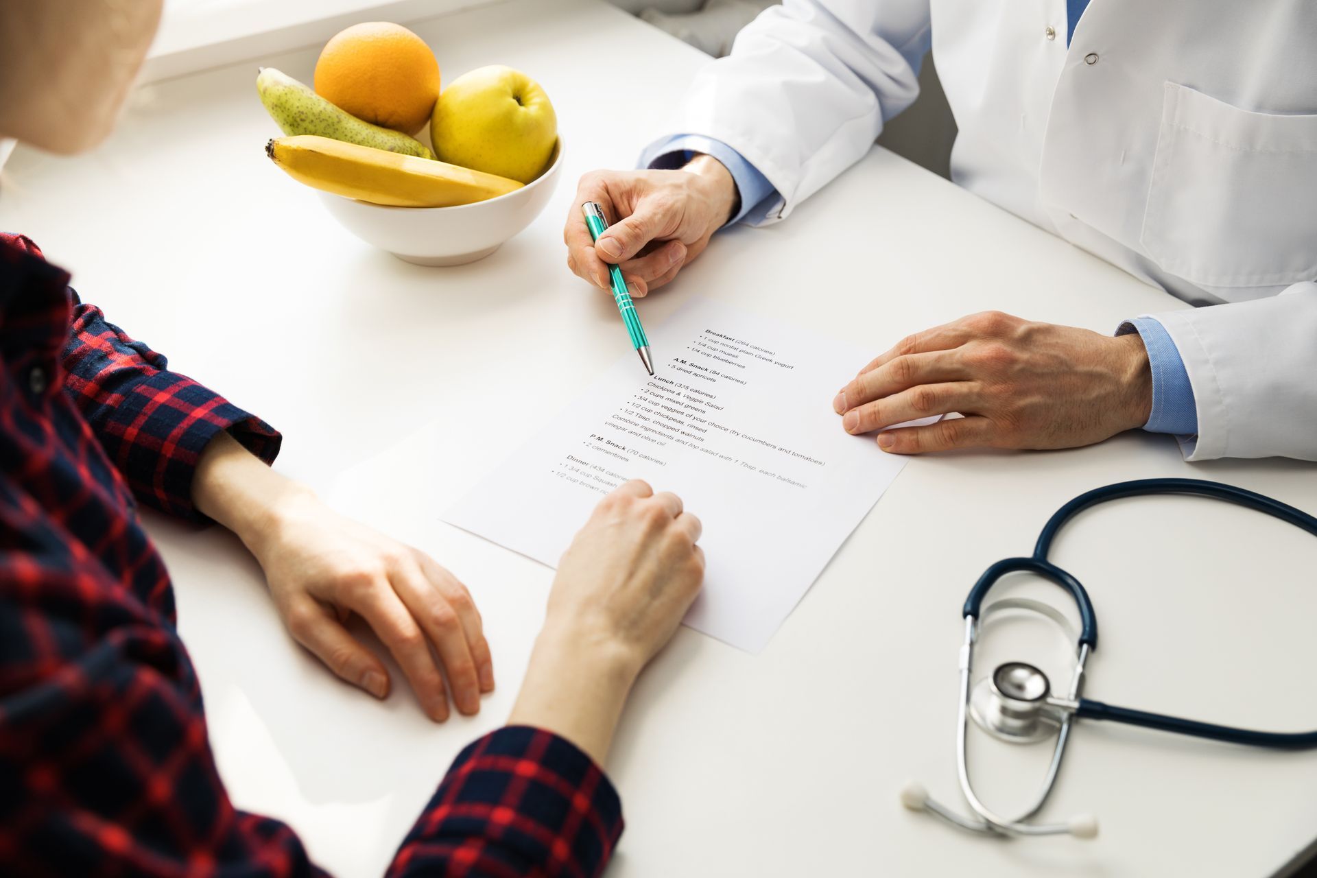 Doctor pointing to document, consulting with patient at a desk. Bowl of fruit and stethoscope present.