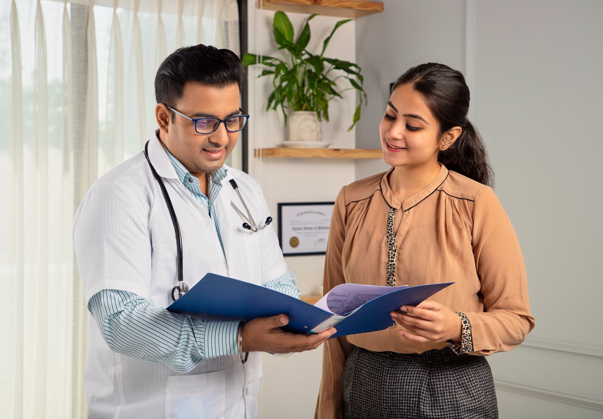 Doctor in a white coat showing a blue folder to a woman in a brown shirt. They are smiling in an office setting.