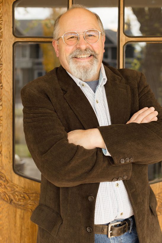 Man with glasses and beard, arms crossed, smiling. Wearing brown blazer, standing by wooden door.