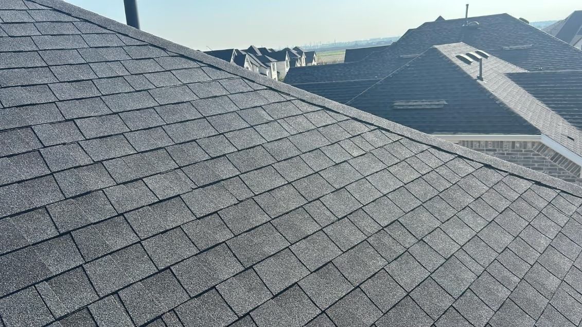 Gray asphalt shingle roof on a house, with other roofs visible in the background on a sunny day.