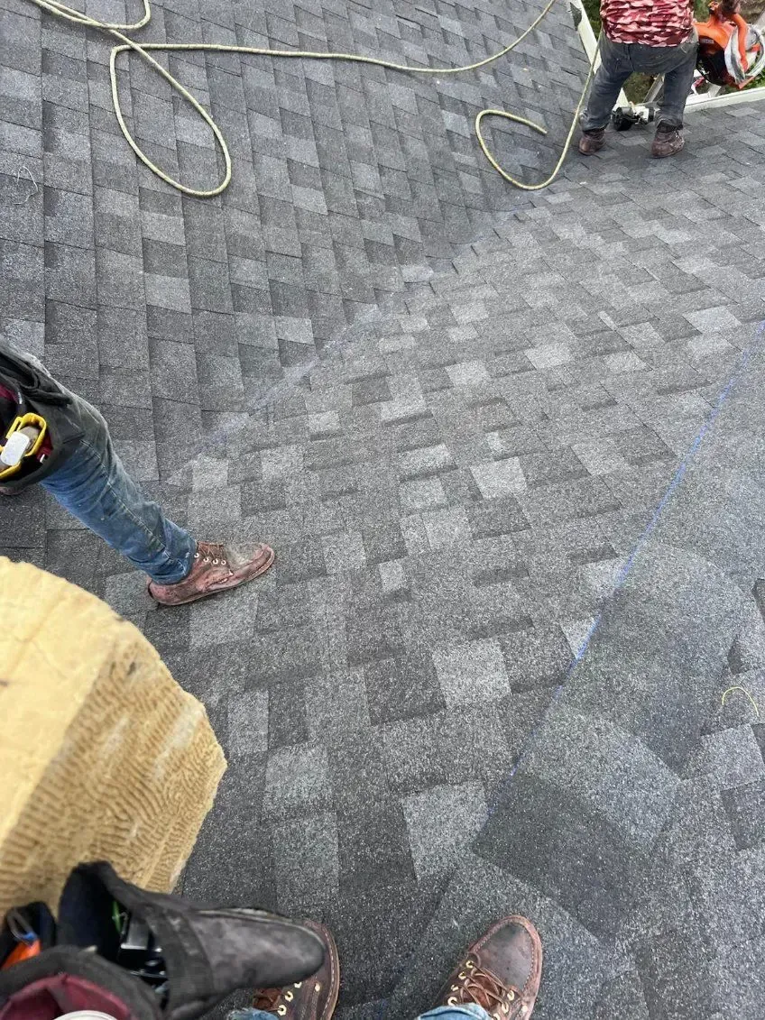People on a gray shingle roof with a rope, one holding a bundle of materials.