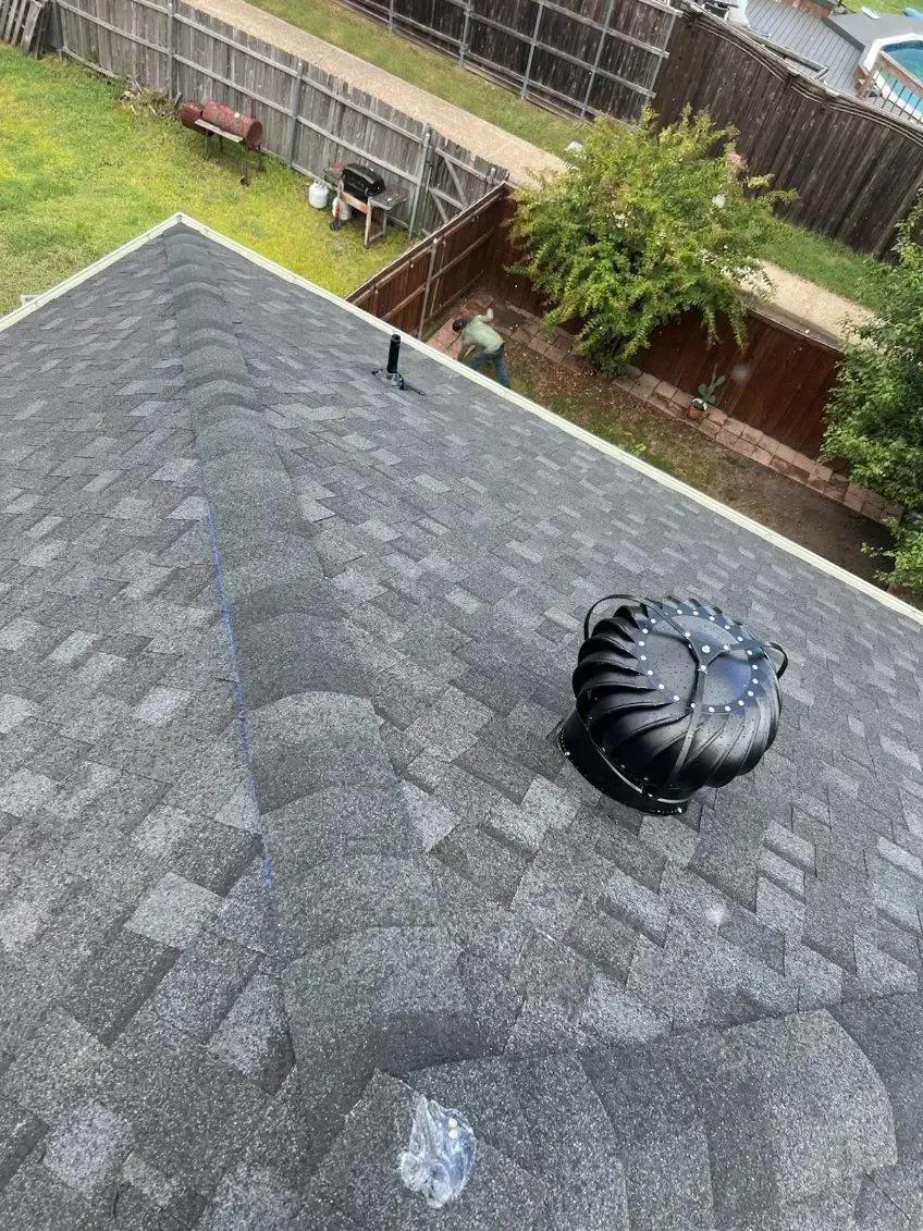 A gray shingle roof with a spinning black turbine vent. Backyard with trees and a fence in the background.