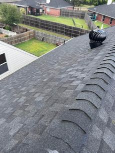 View of a dark shingle roof with a rotating vent, with a backyard and other houses in the background.