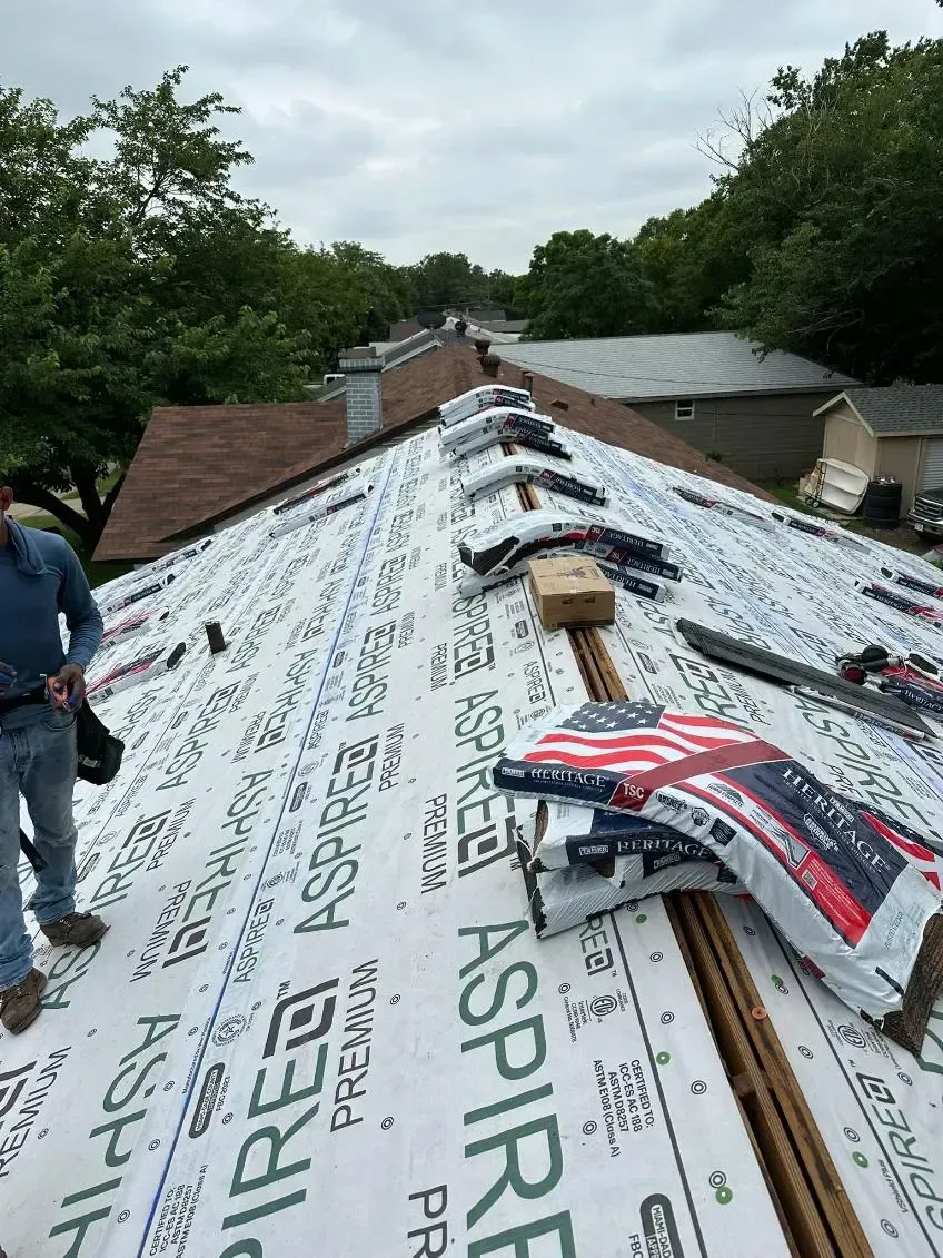 Roofing work in progress; person on roof, laying down underlayment; houses and trees in background.