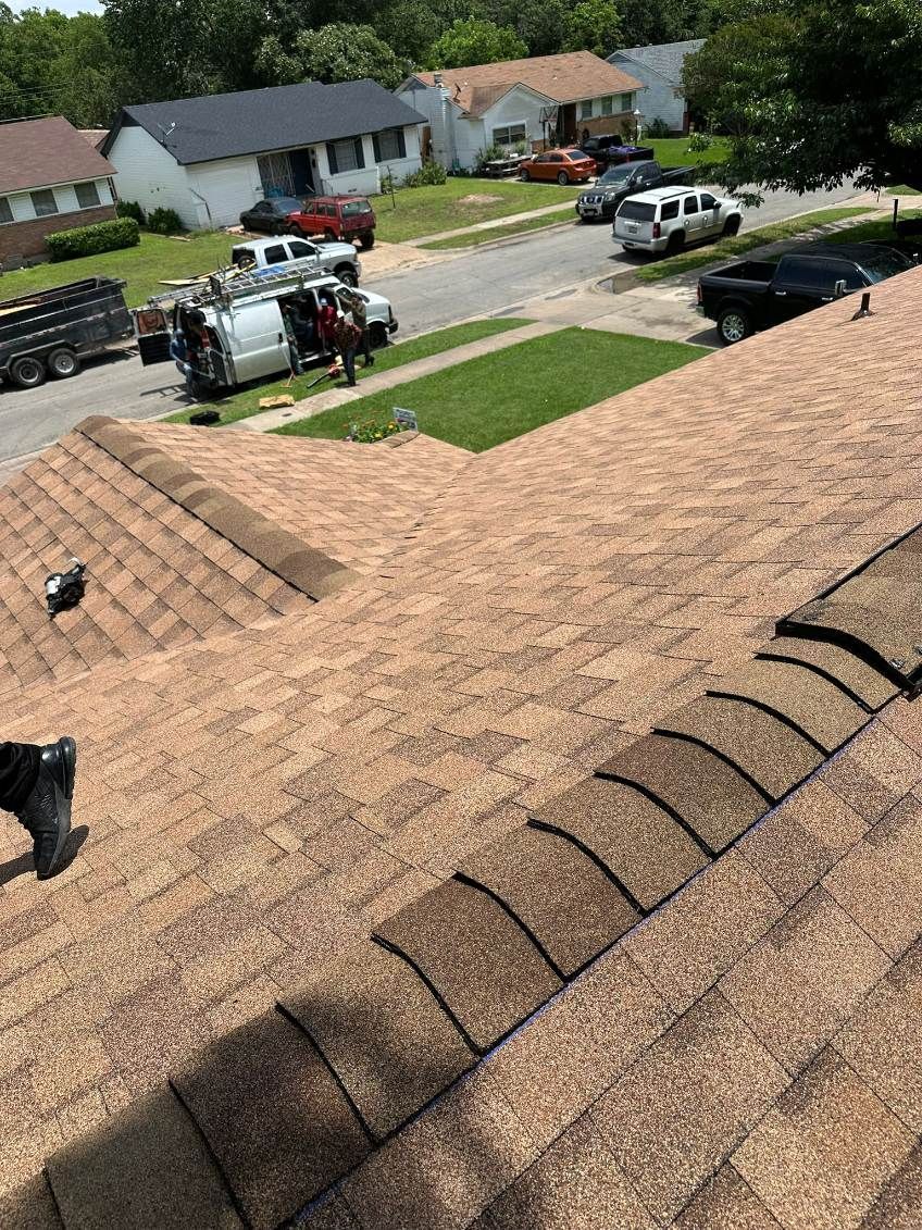 Roofer on a brown shingle roof; crew, van, and neighborhood street visible in background.