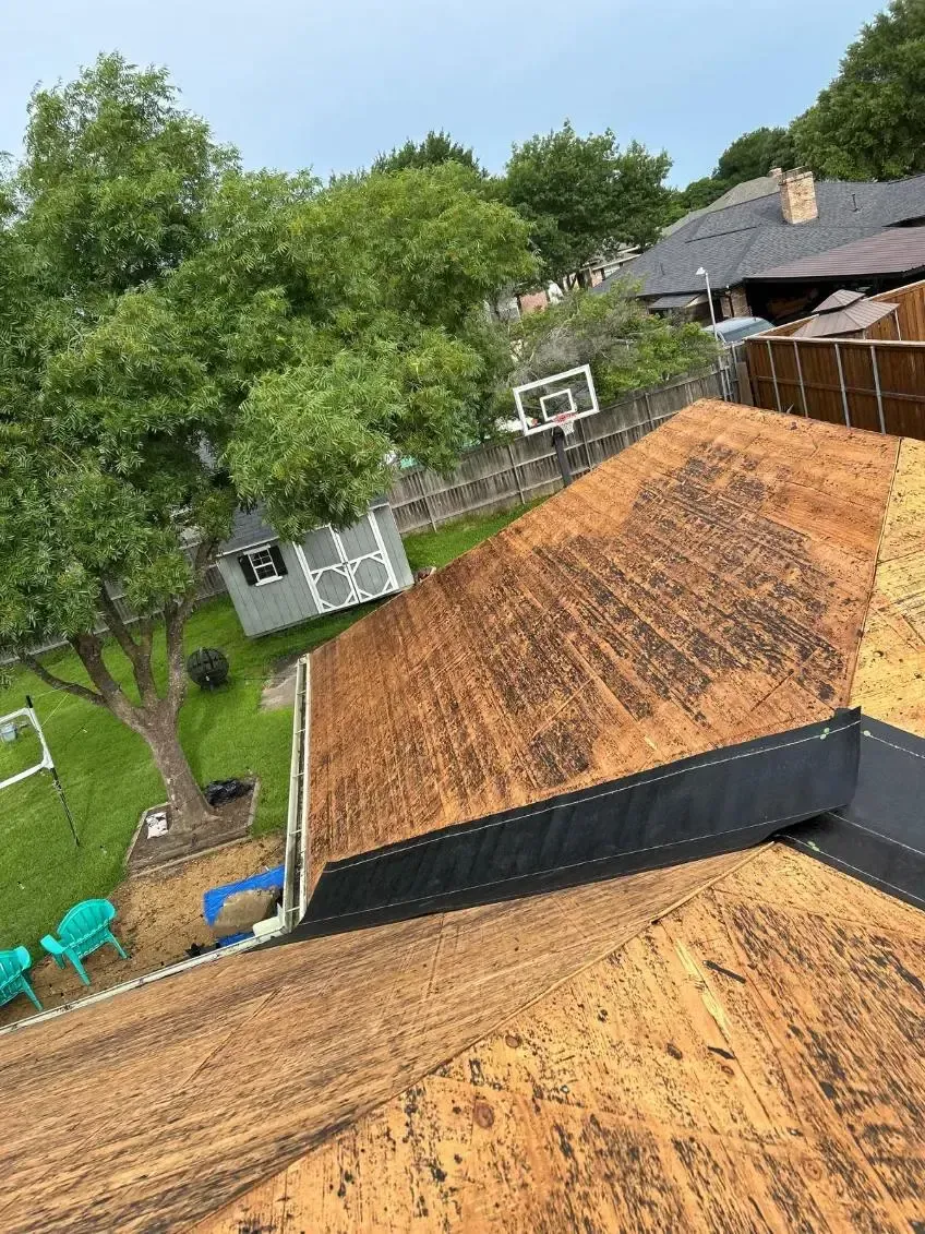 Roof partially covered with wood and dark material, backyard with trees and houses in the background.
