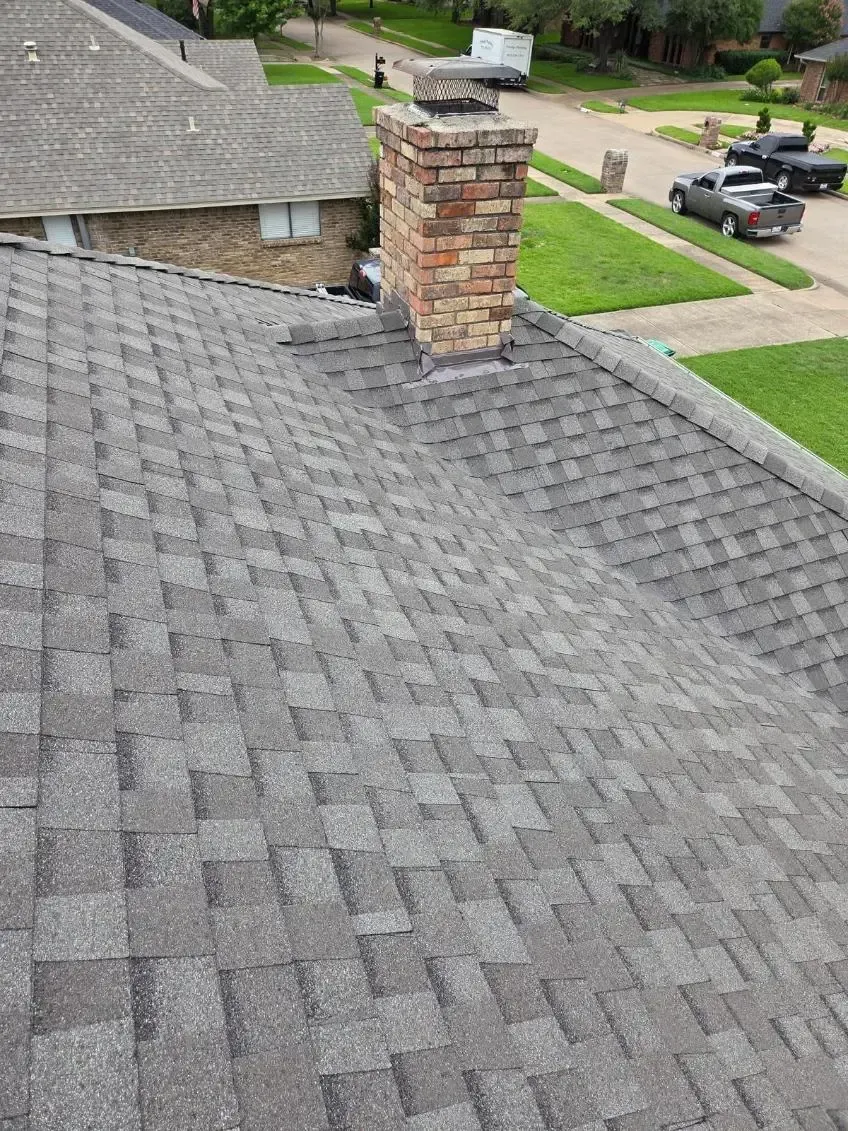 Gray shingle roof with a brick chimney. Green lawn and parked cars visible in the background.