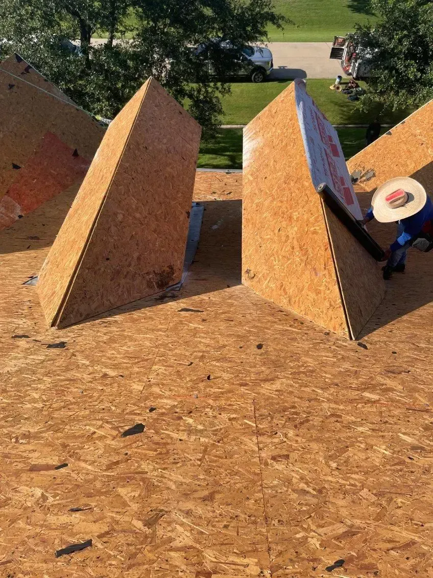Construction workers on a roof, installing angled plywood pieces, with a blue shirt and a white car in the background.