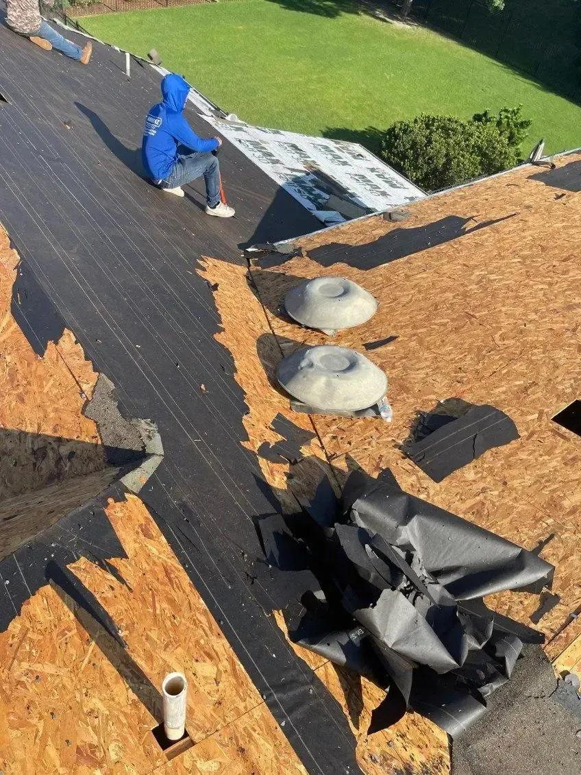 Person on a roof removing old roofing material. Exposed wooden surface and vents are visible.