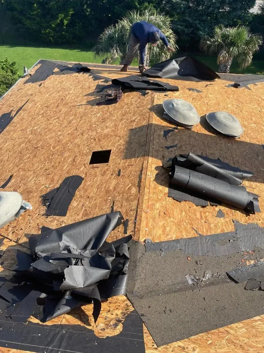 Person removing old roofing material from an OSB roof deck. Sunlight, three vents, and dark underlayment are visible.