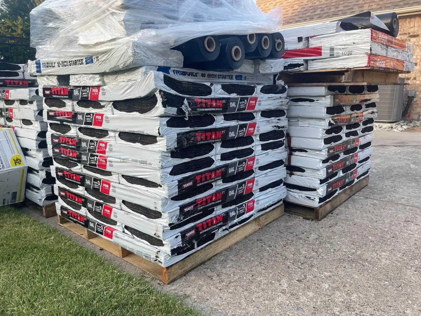 Pallets of roofing materials stacked outdoors, wrapped in plastic.