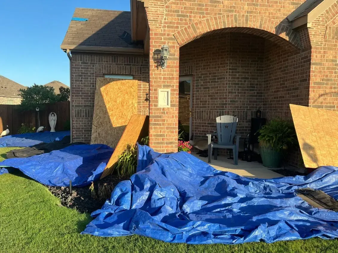 Blue tarps cover lawn near a brick house with damaged roof and exposed wood panels.