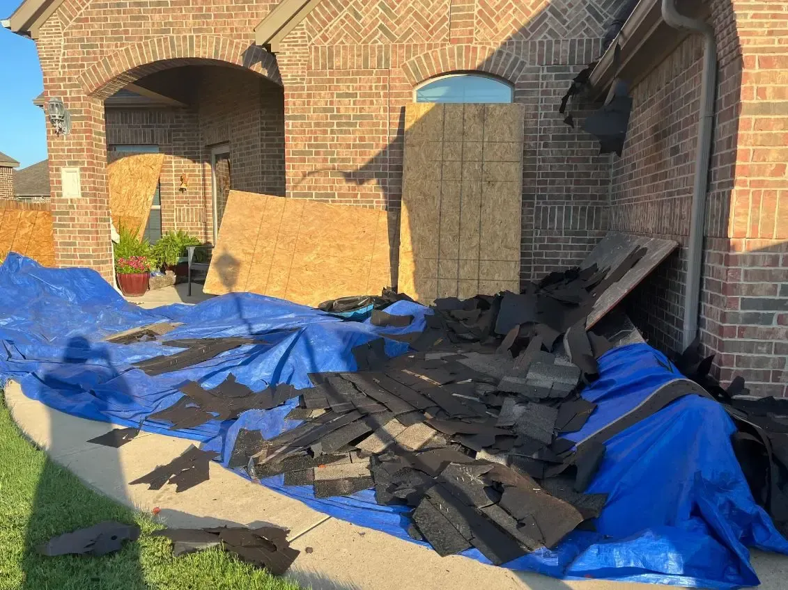 Brick house with roof damage, plywood covering a section, debris on a blue tarp on the lawn.