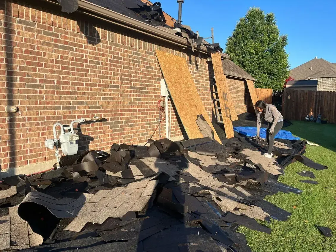 Damaged roof with fallen shingles near a brick house; a person is on the grass.