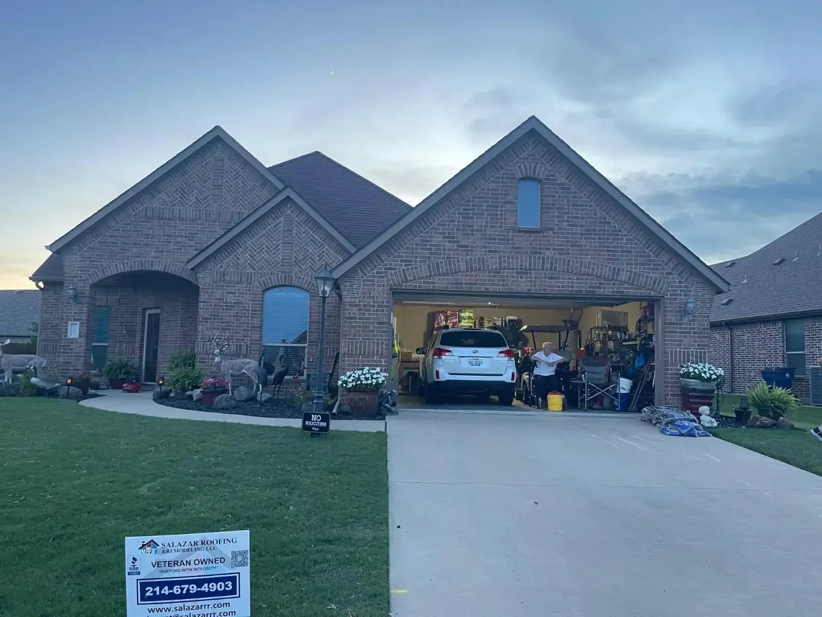 Brick house with open garage, driveway, and lawn. A car is parked inside the garage. People are visible.