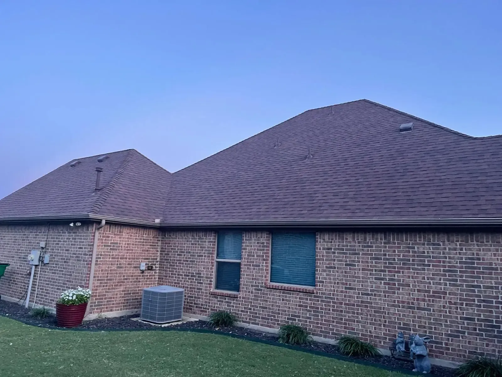 Brick house with brown roof, blue sky background. Green grass and landscaping in front.