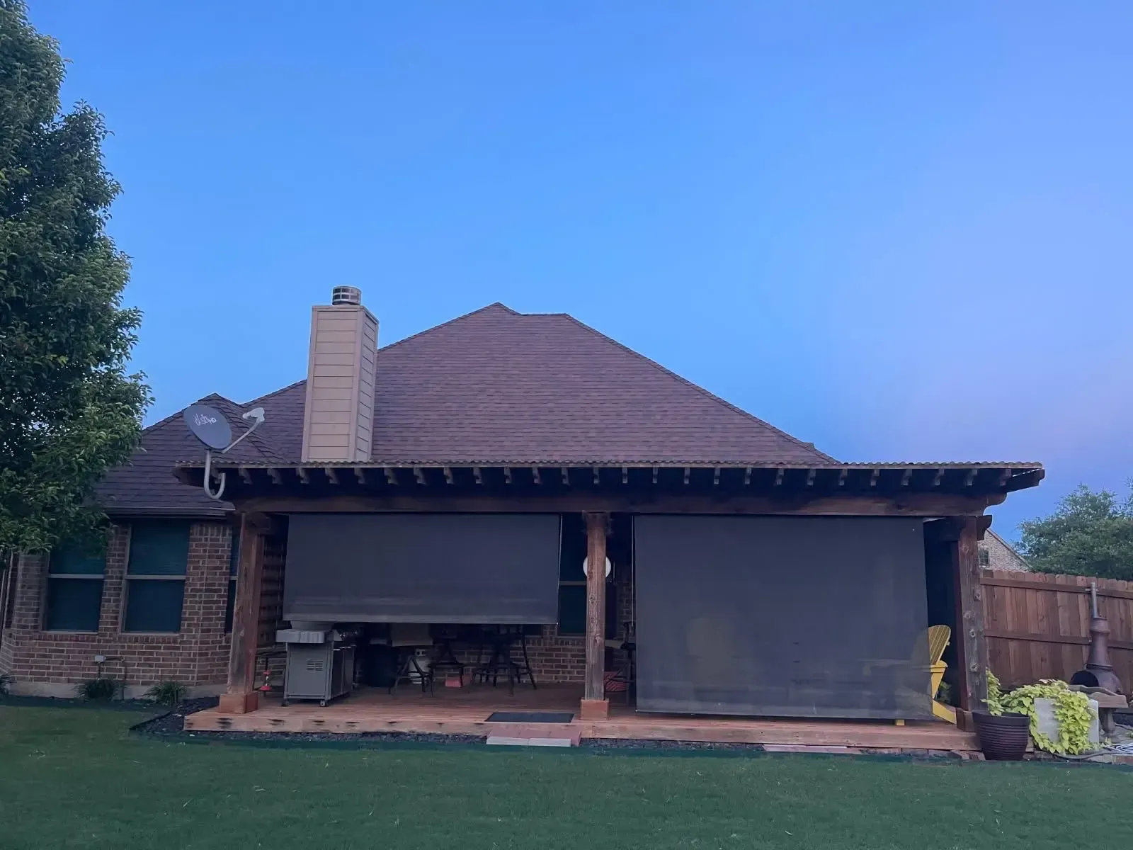 Outdoor patio with shades, attached to a brick house, green yard, blue sky.