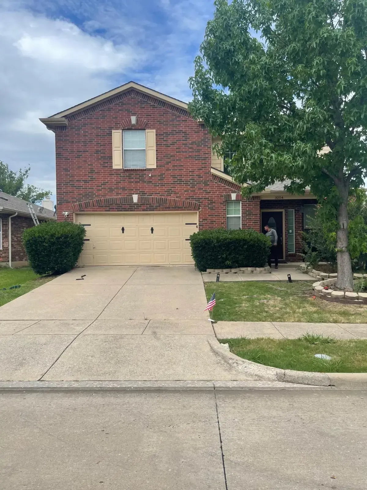 Two-story brick house with a tan garage door and a concrete driveway. A person stands at the front door.