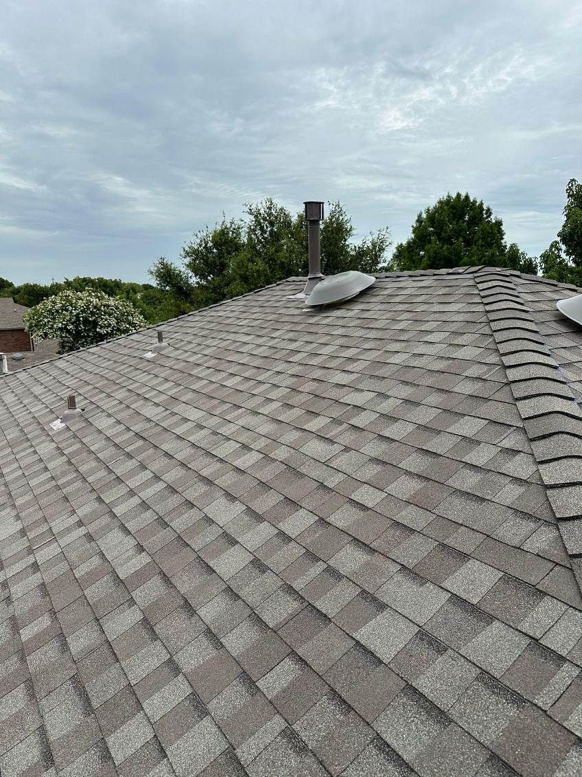 A brown shingle roof with a chimney and vents, set against a cloudy sky.