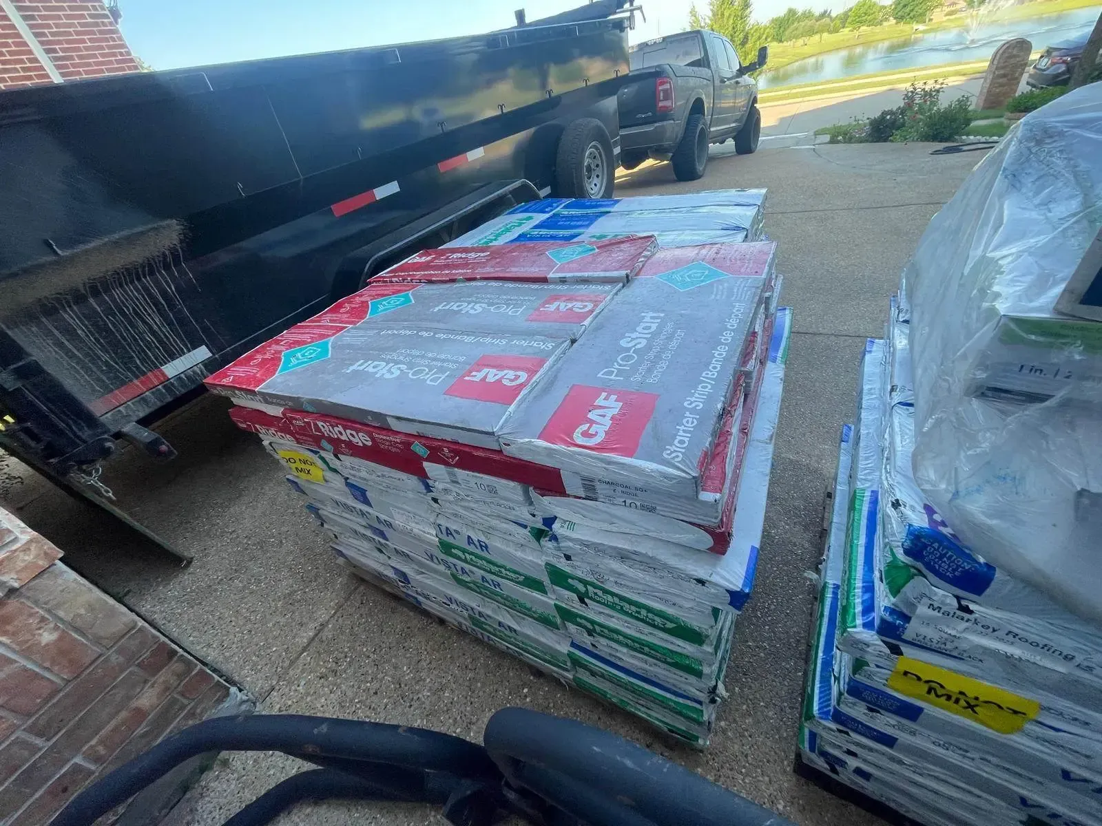 A stack of roofing shingles in various colors next to a trailer and truck outdoors.