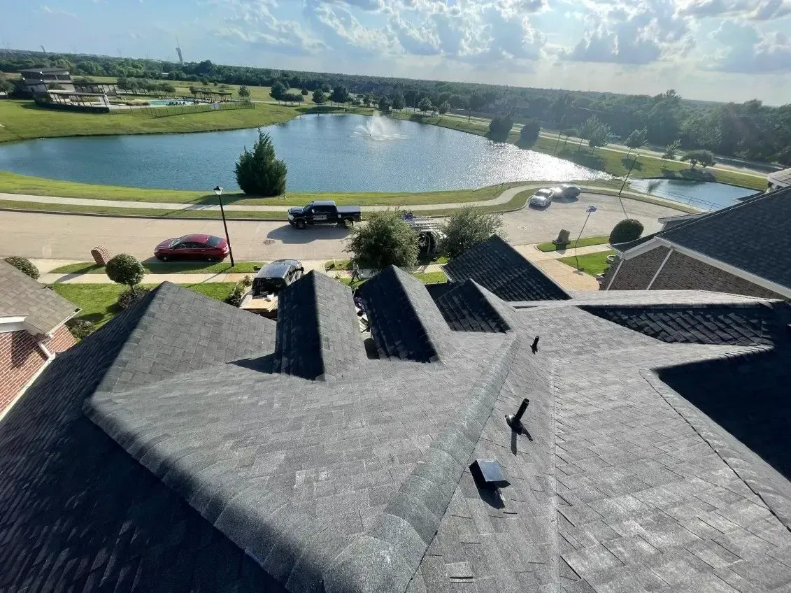 View of a dark shingled roof with a lake and road in the background, under a sunny sky.