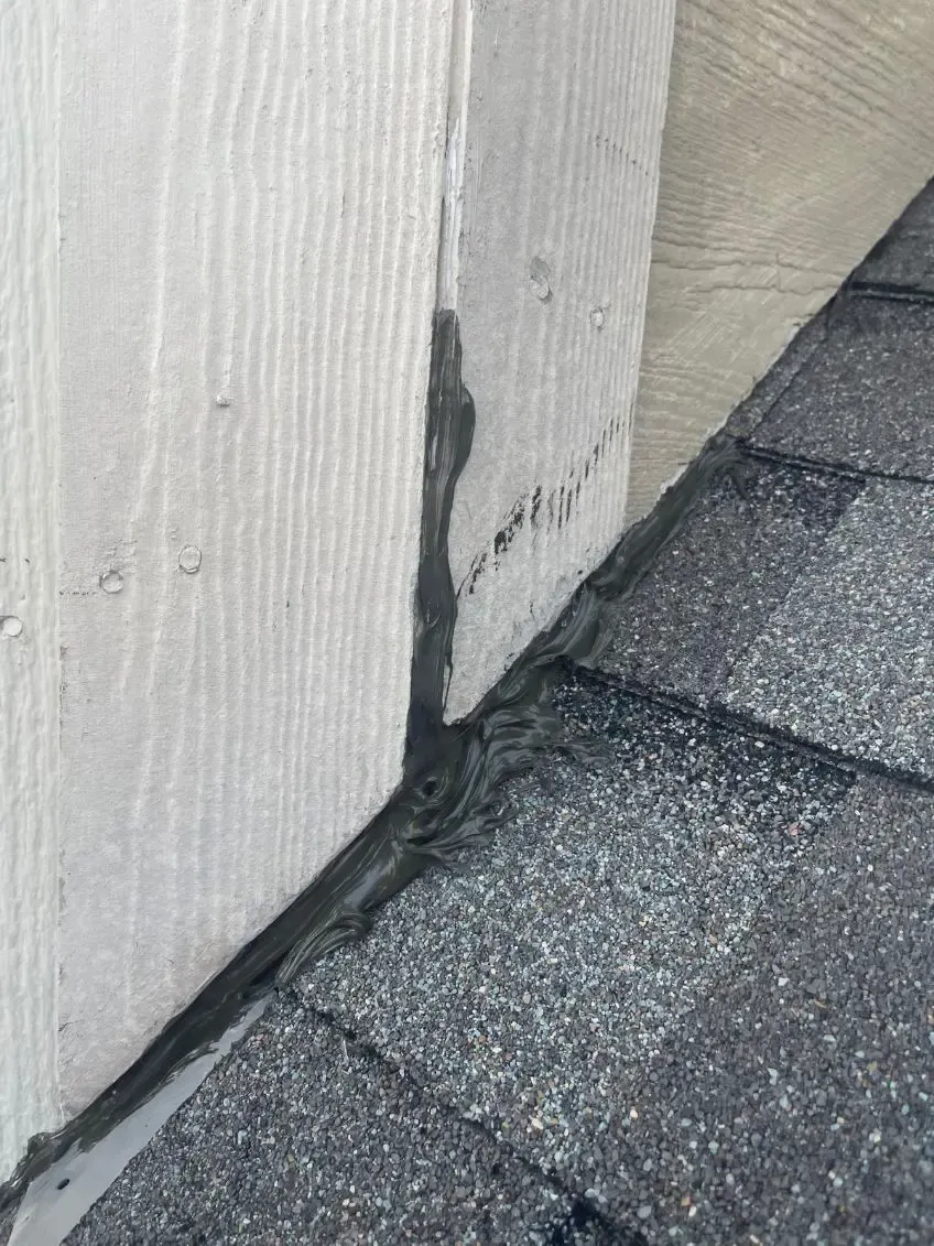Gray roof shingles meeting a white wall, sealed with black caulk.
