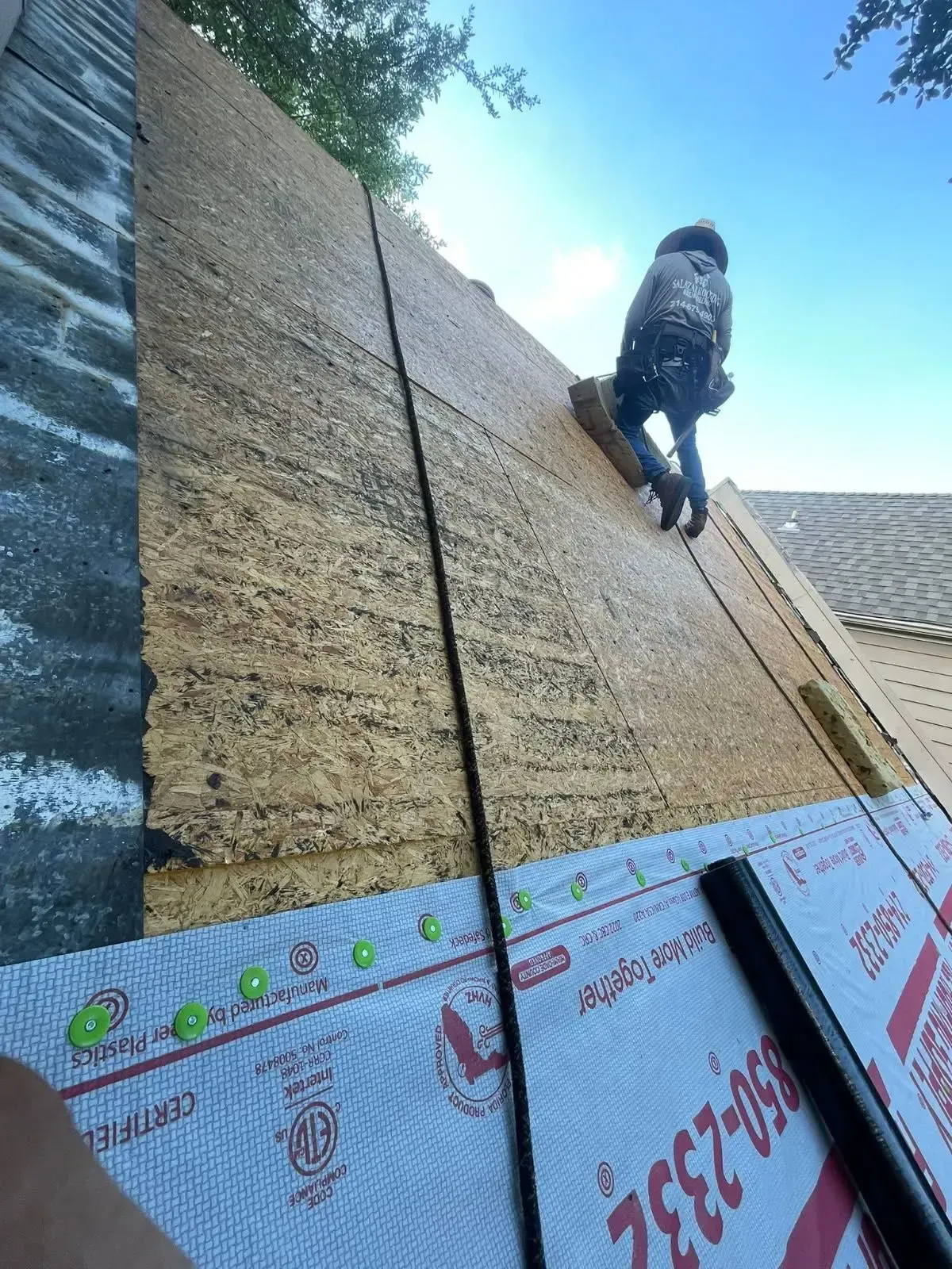 Roofer working on a roof, secured by a safety rope. New underlayment visible. Sunny day.