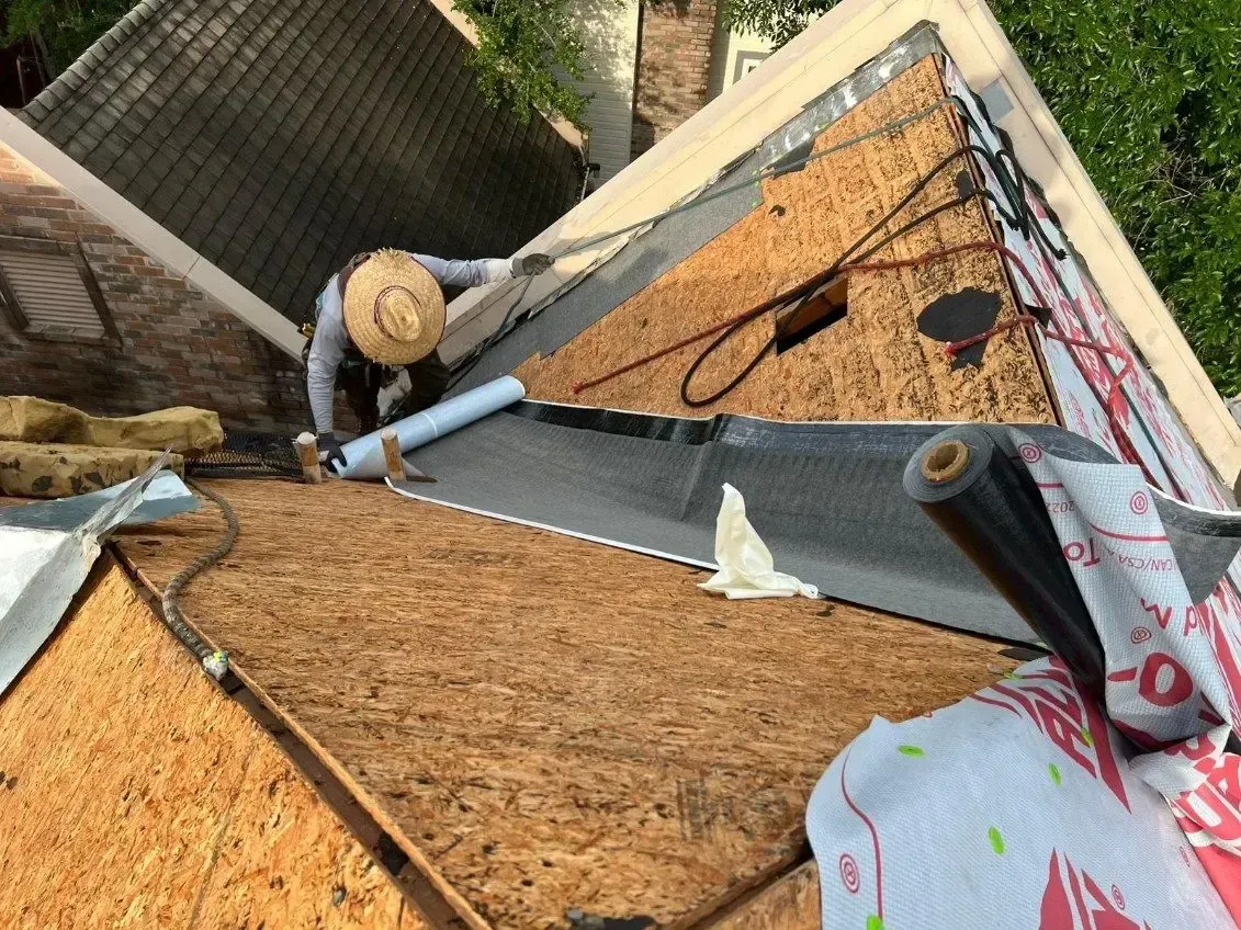 Roofer in straw hat installing black roofing material on a wooden roof.