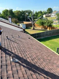View of a brown shingled roof with several vents. A person stands near the edge with a neighborhood in the background.