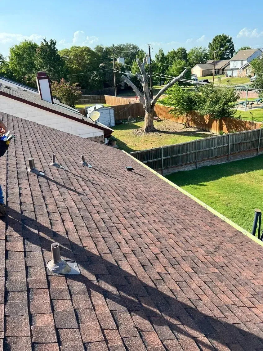 Brown shingled roof with vent pipes, viewed from above, with residential landscape in background.