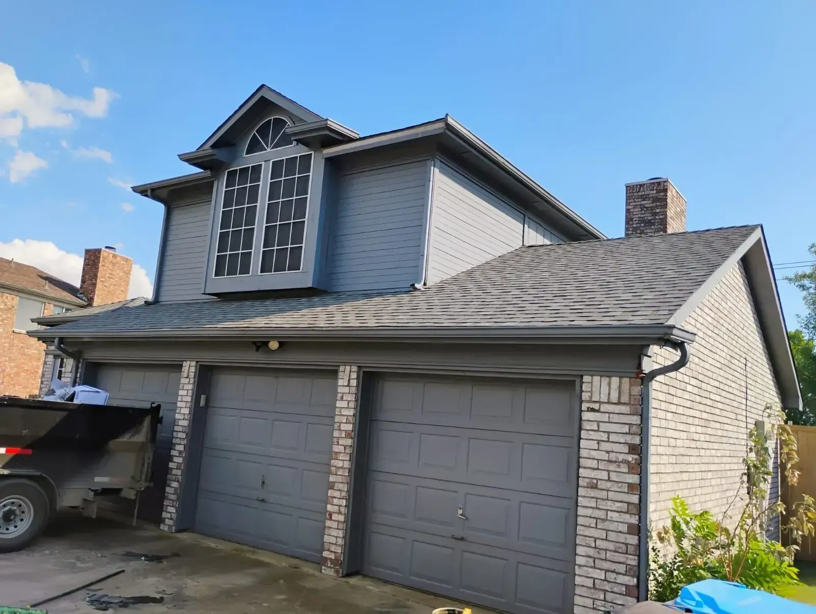 Two-story gray house with brick accents, three garage doors, and a chimney against a blue sky.