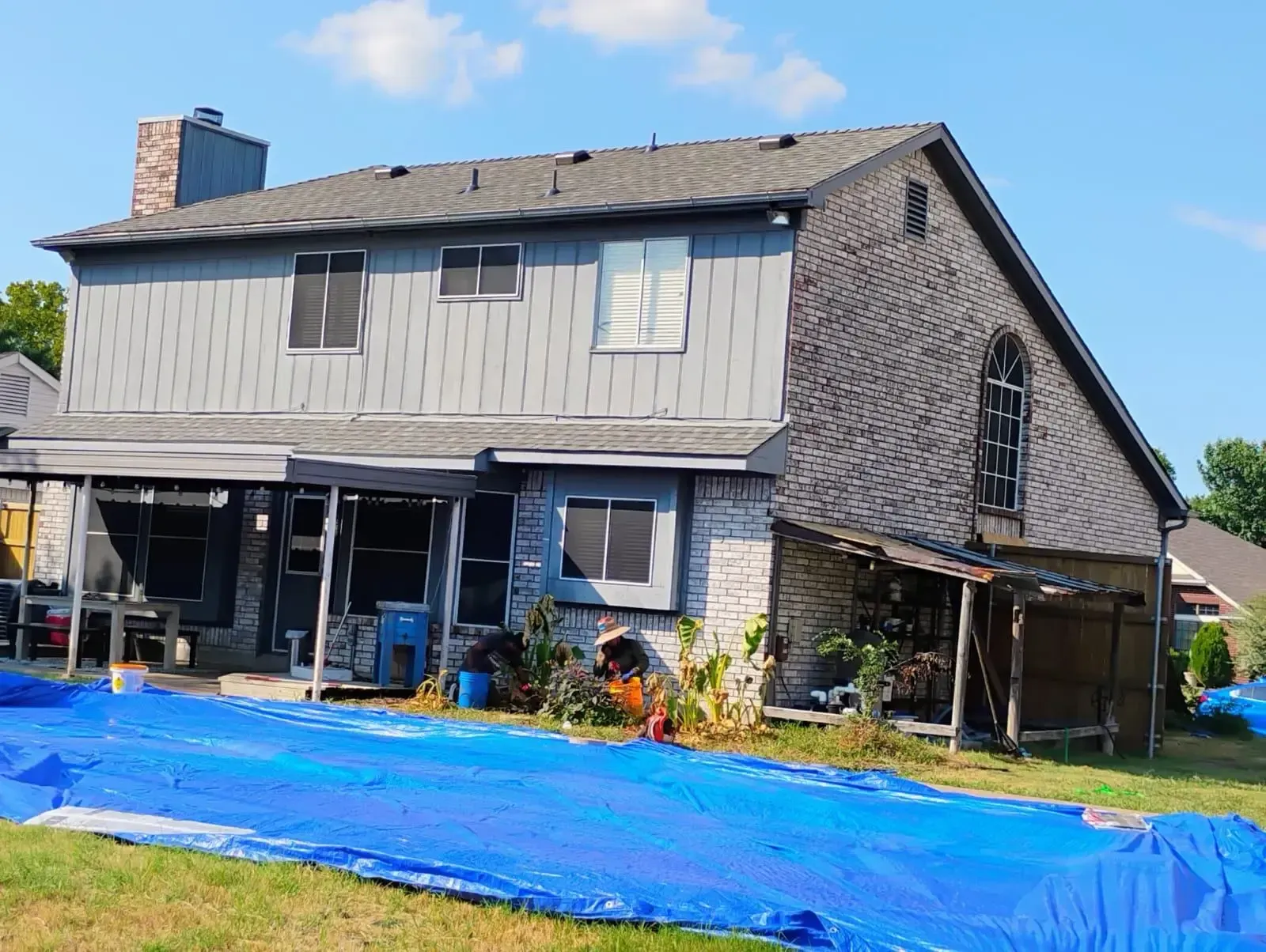 Back of a two-story house, gray siding, stone facade, blue tarp in yard, roof damage.