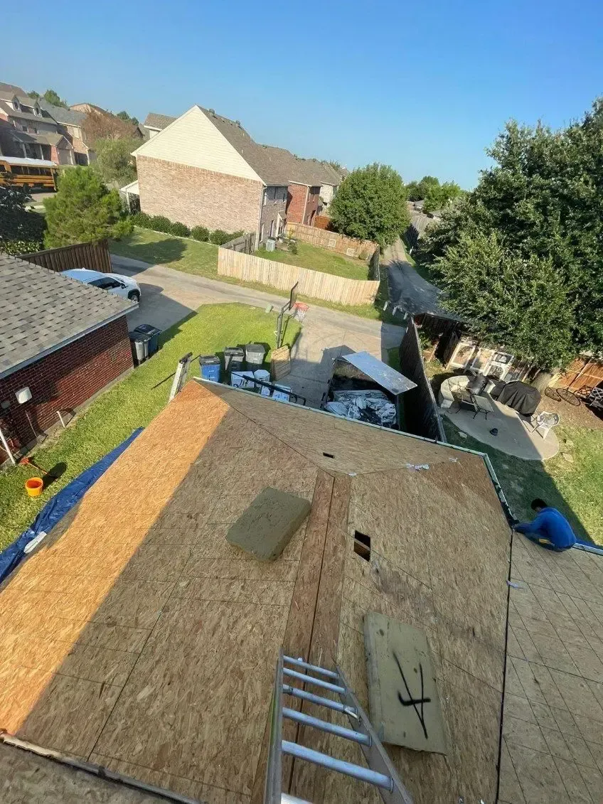 Roofer working on a partially shingled roof with a ladder, blue skies, and houses in the background.