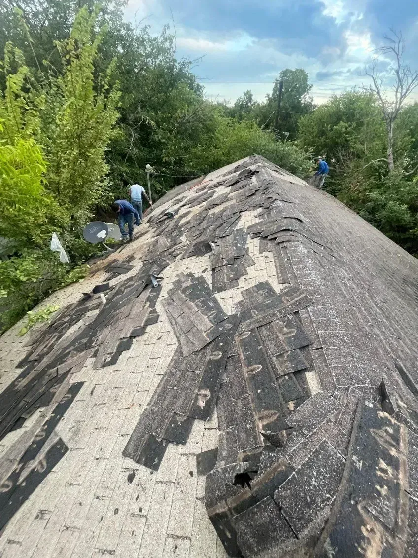 Workers on a weathered rooftop, removing damaged shingles. Surrounded by green trees under a cloudy sky.