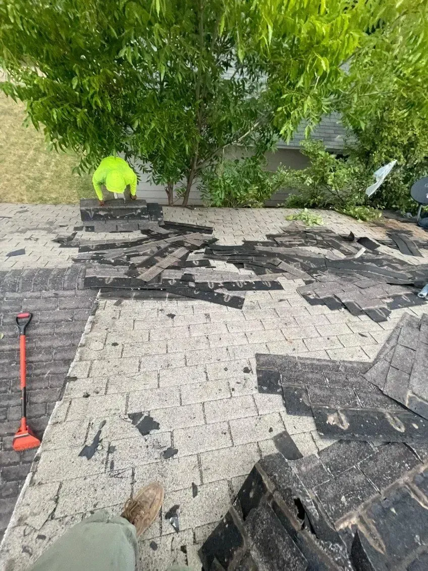 Roofer in neon shirt removes asphalt shingles from a roof. Shingles are scattered; shovel nearby.