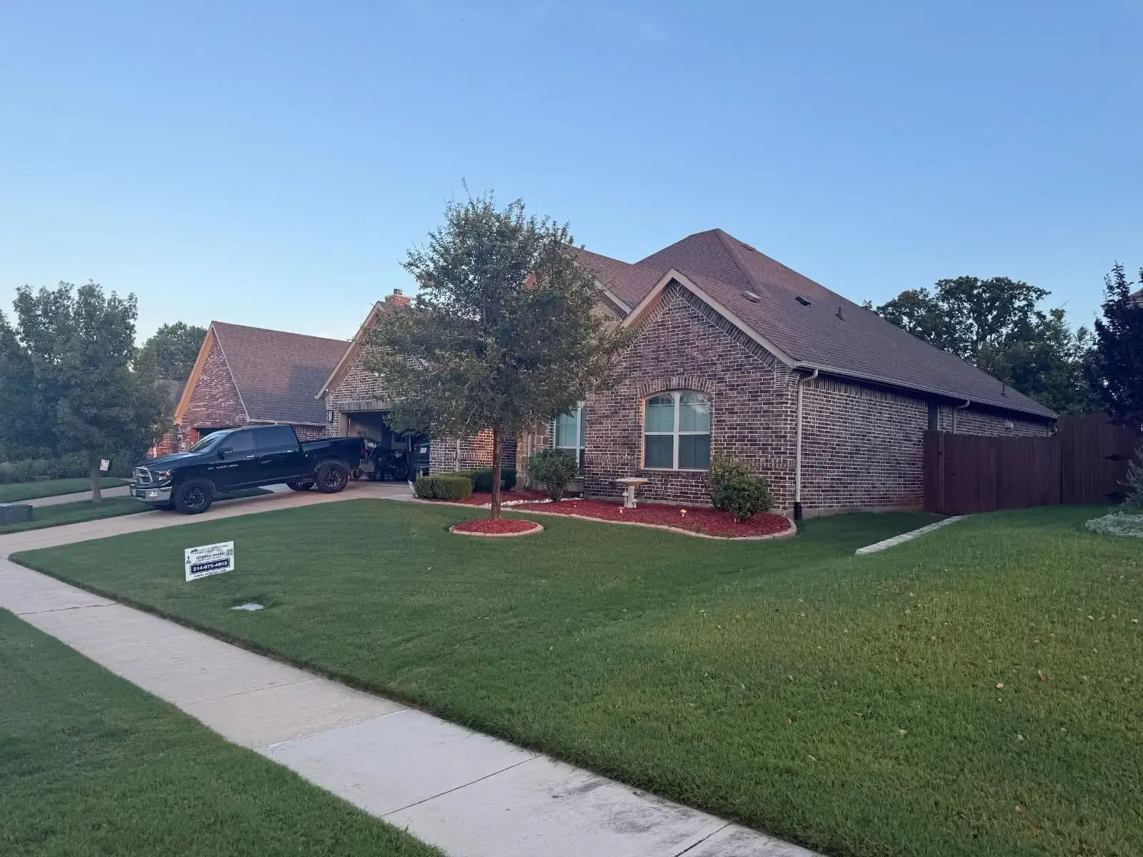 A brick house with a dark truck in the driveway. Green lawn and trees in a residential setting.