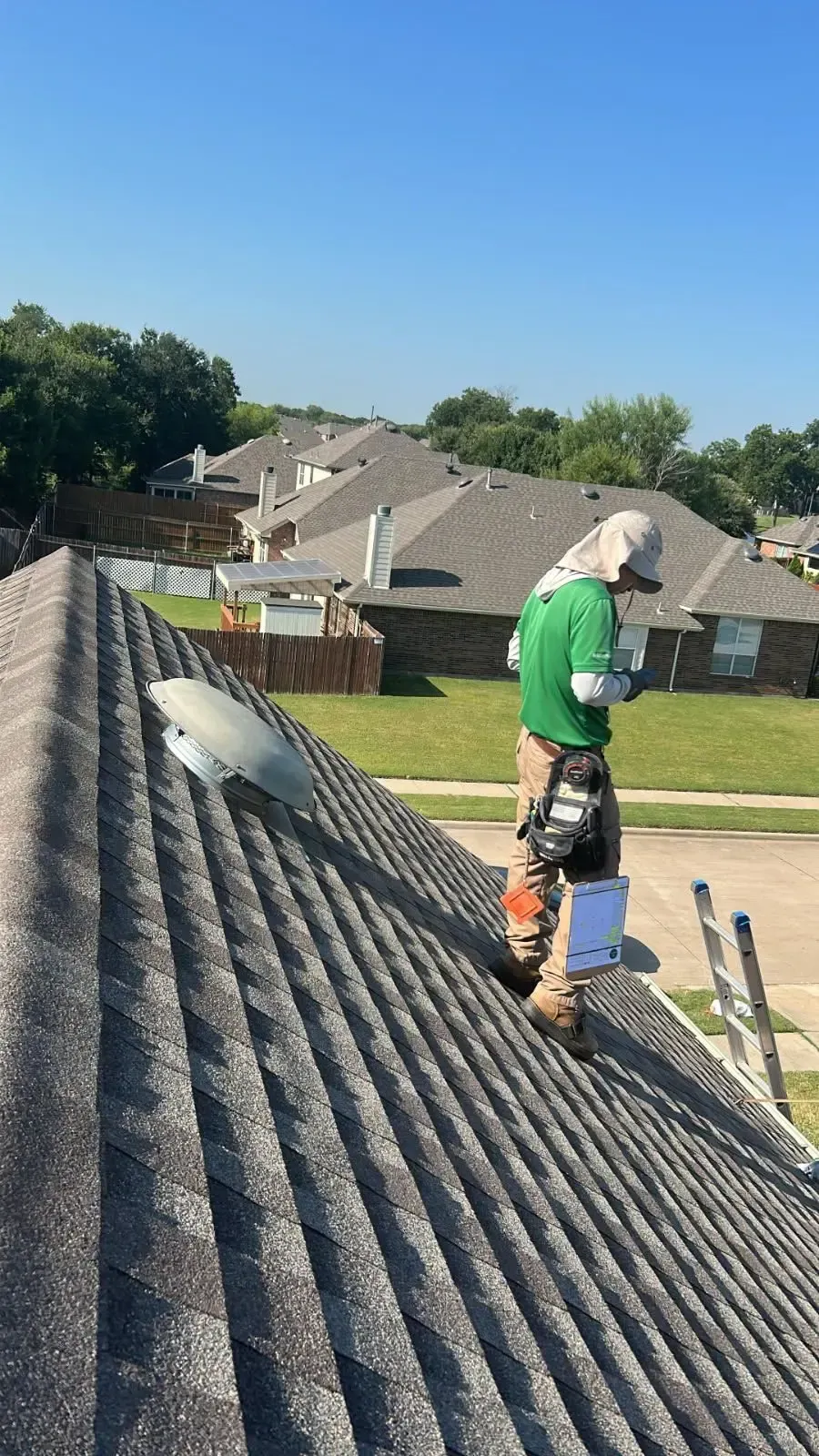 Roofer in green shirt on a gray shingle roof, holding clipboard. Blue sky, suburban setting.