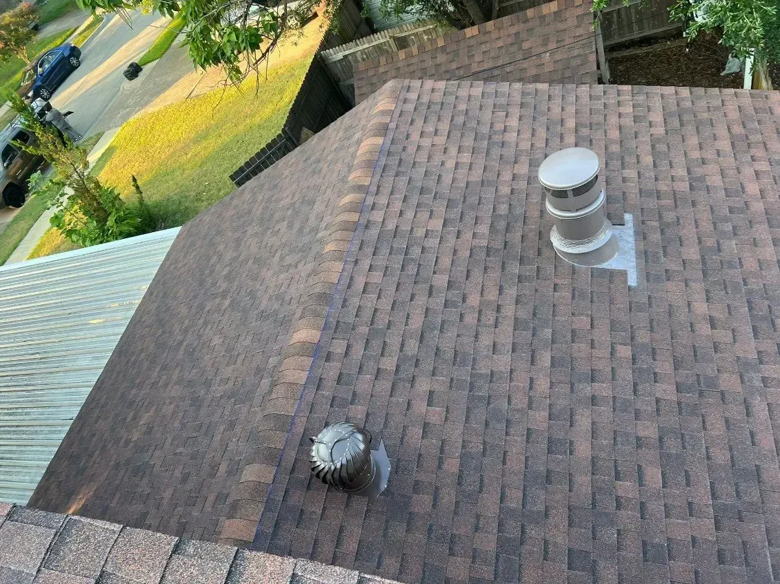 Overhead view of a brown asphalt shingle roof with a vent pipe and chimney. Green grass and a road are visible.