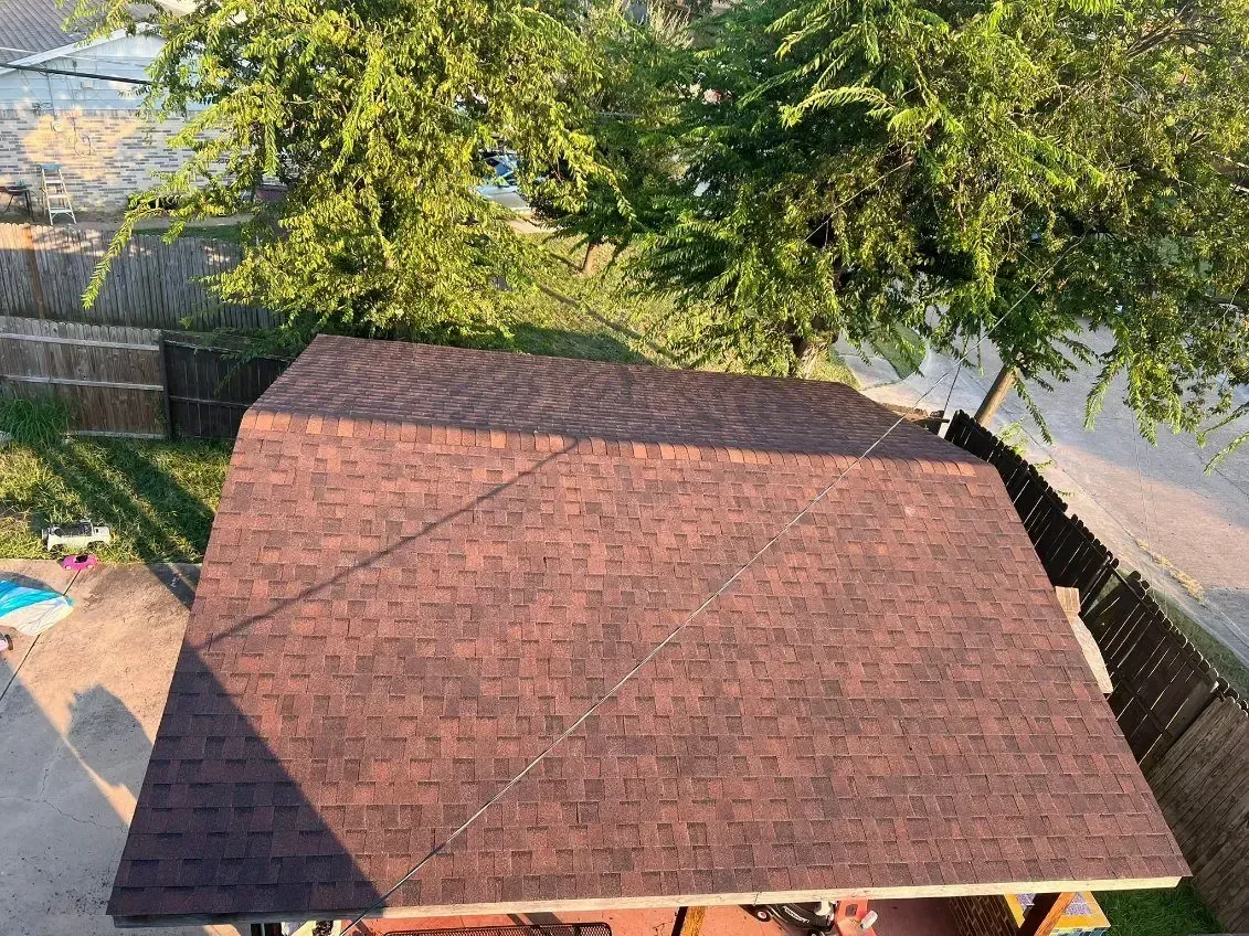 Brown shingled roof of a structure, viewed from above, with surrounding trees and a fence.