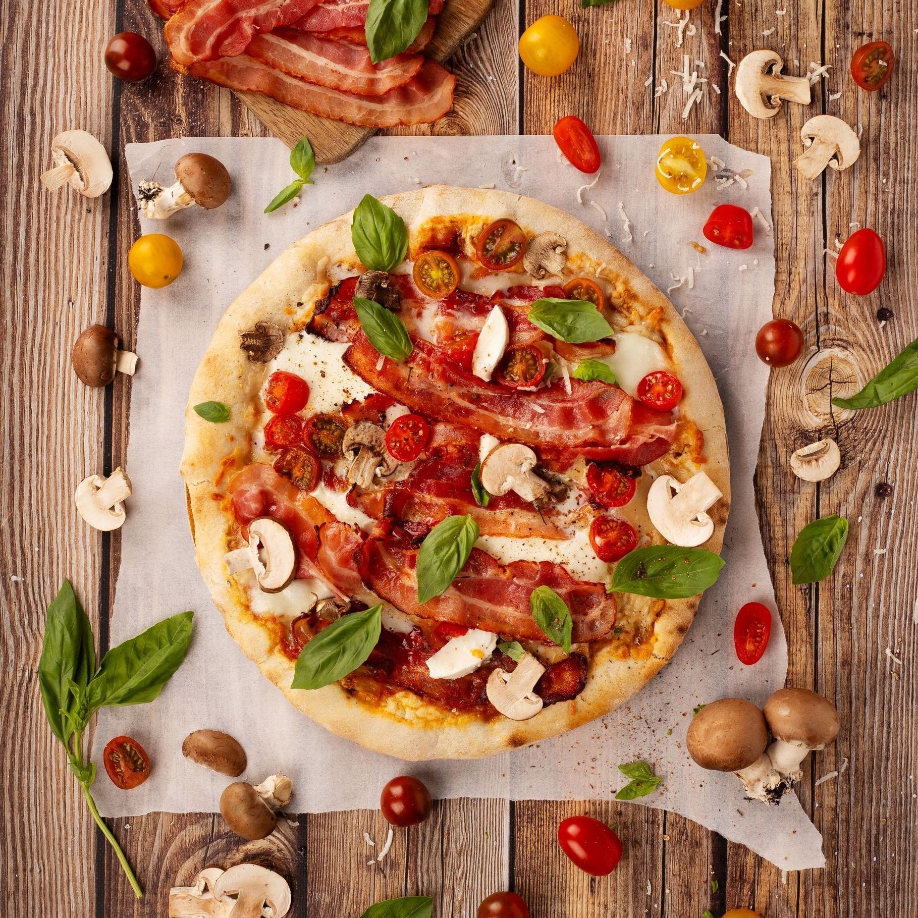 Une pizza avec bacon, champignons, tomates et basilic sur une table en bois.