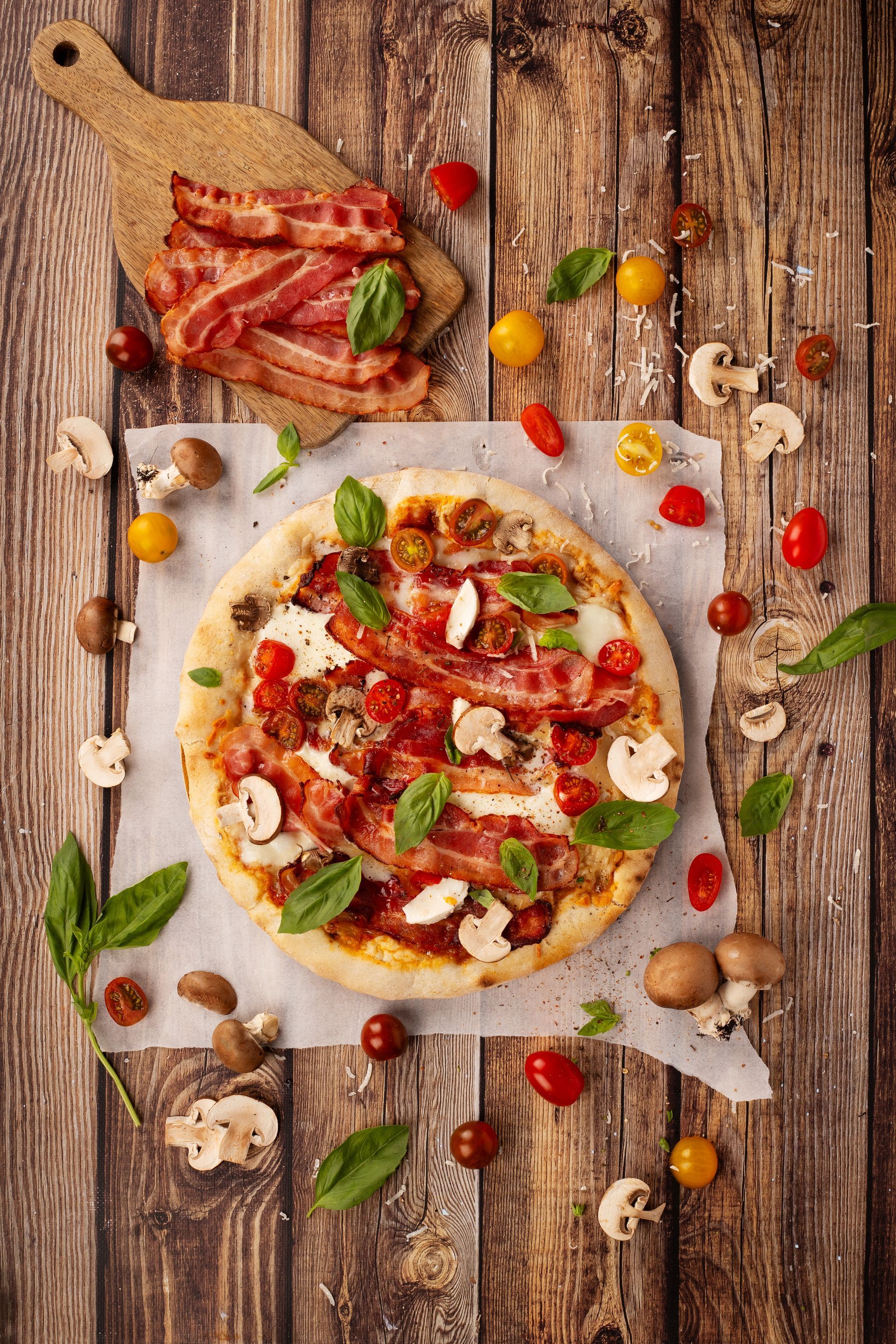 Une pizza avec tranches de crispy bacon, champignons, tomates et basilic sur une table en bois.