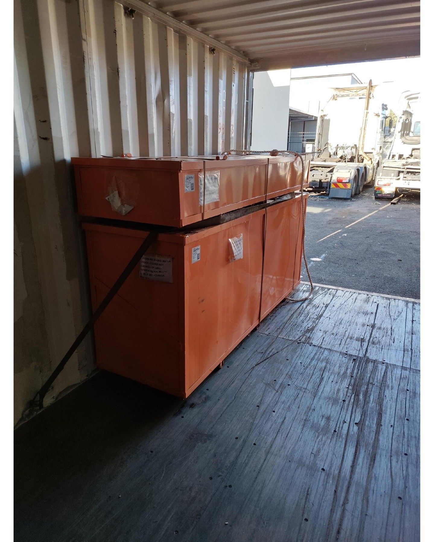 Orange boxes in a shipping container, secured with straps. Background shows vehicles and outdoor setting.