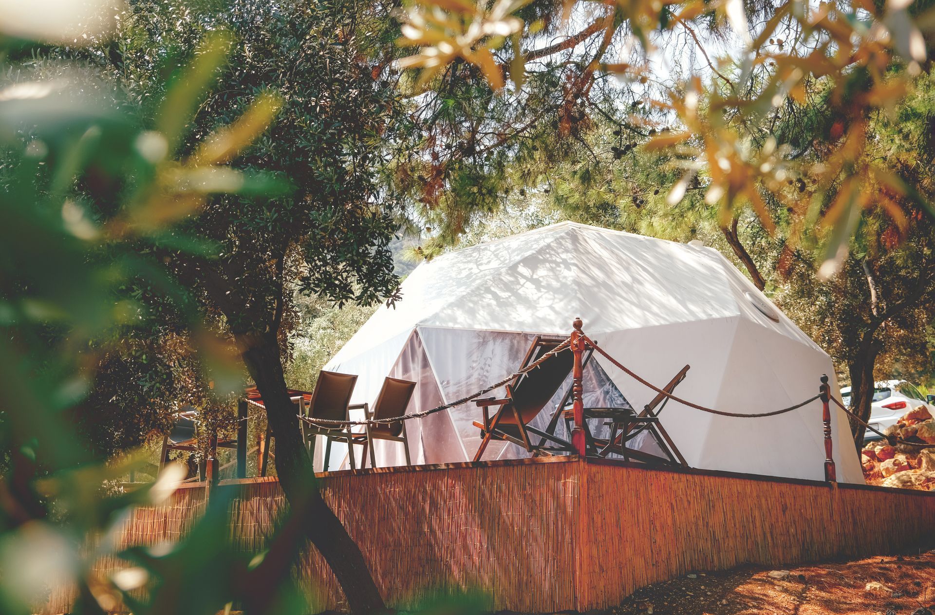White dome tent on a wooden platform surrounded by trees, with chairs outside.