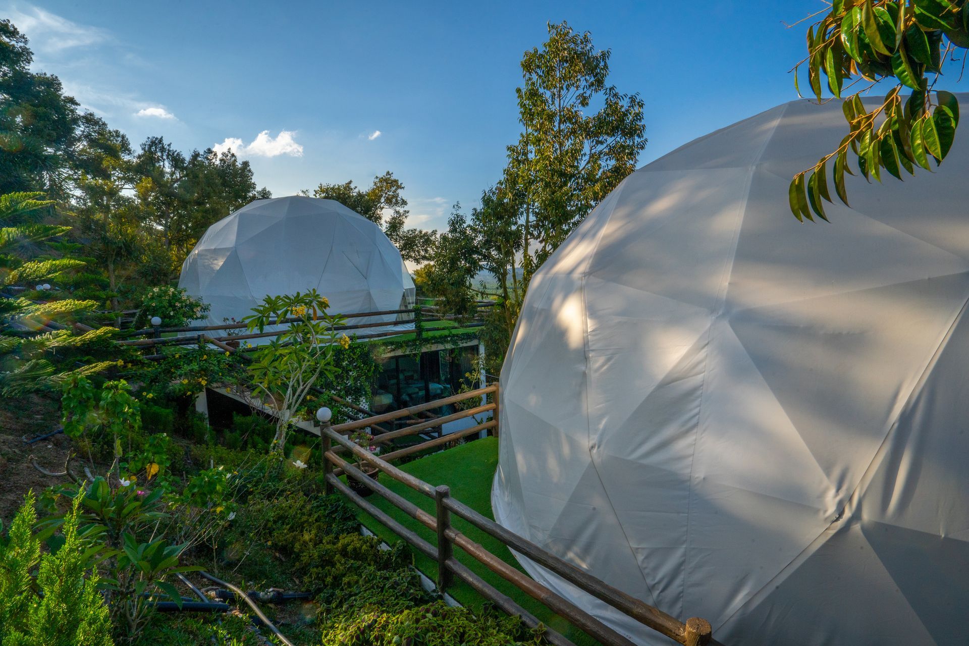 Two white dome-shaped structures on a hillside, connected by a wooden walkway, surrounded by greenery and trees.