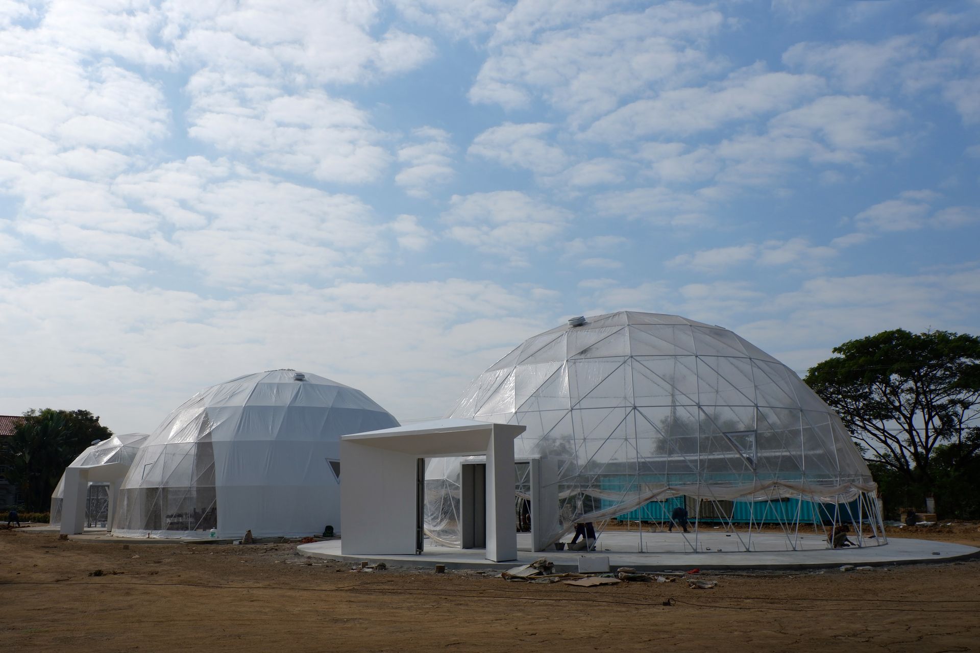 Three white geodesic domes under construction on a dirt lot, against a blue sky.