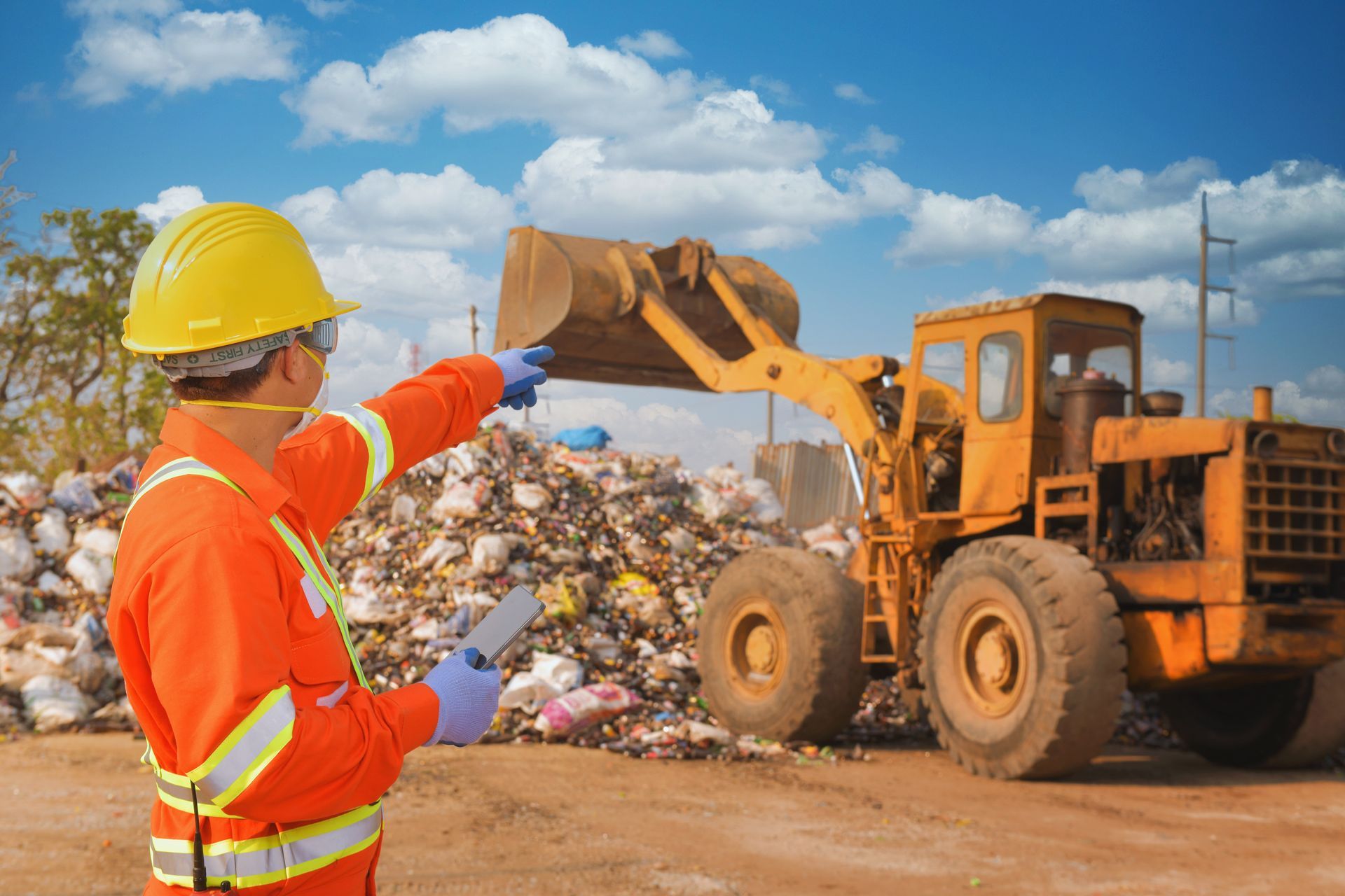 Worker in orange safety gear directs a yellow loader at a landfill under a blue sky.