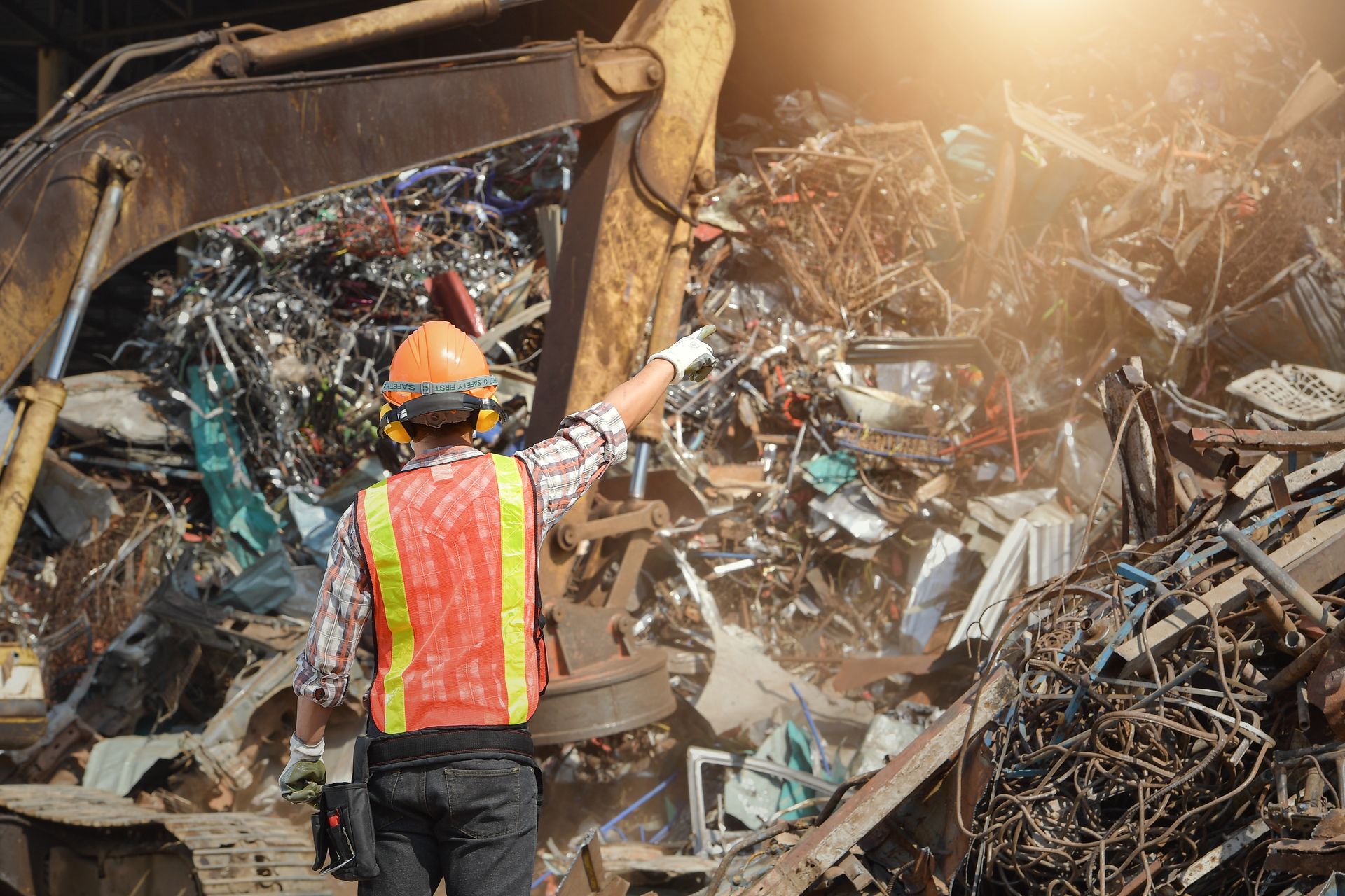 Worker in hard hat and safety vest pointing at a large pile of scrap metal at a recycling yard Worker in hard hat and safety vest pointing at a large pile of scrap metal at a recycling yard
