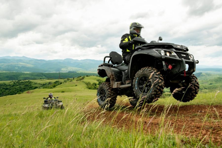 Person on ATV jumping over a dirt path in a grassy, mountainous landscape. Another ATV in the background.