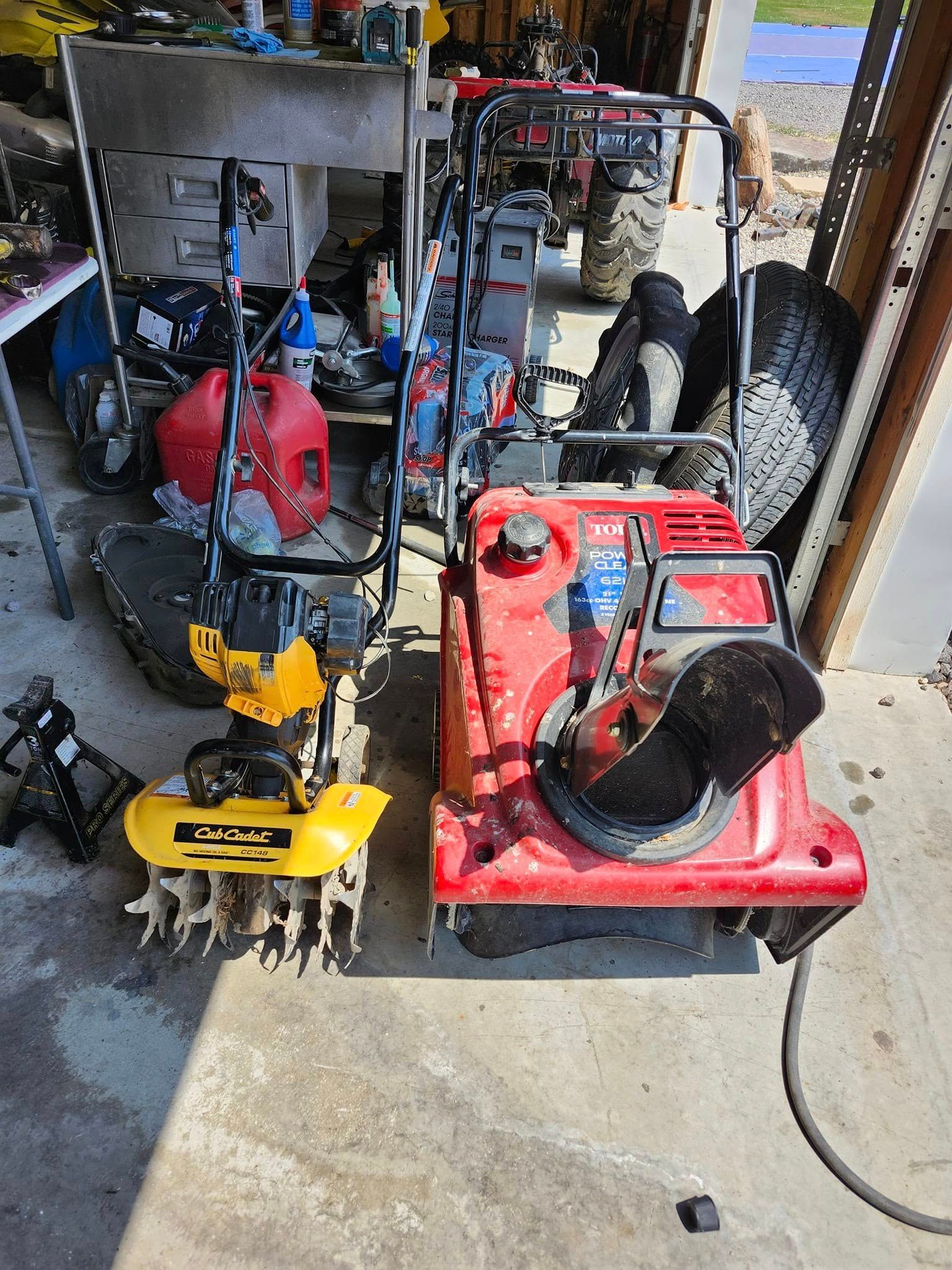 Two pieces of equipment: a yellow tiller and a red snow blower, sit side by side in a garage setting.