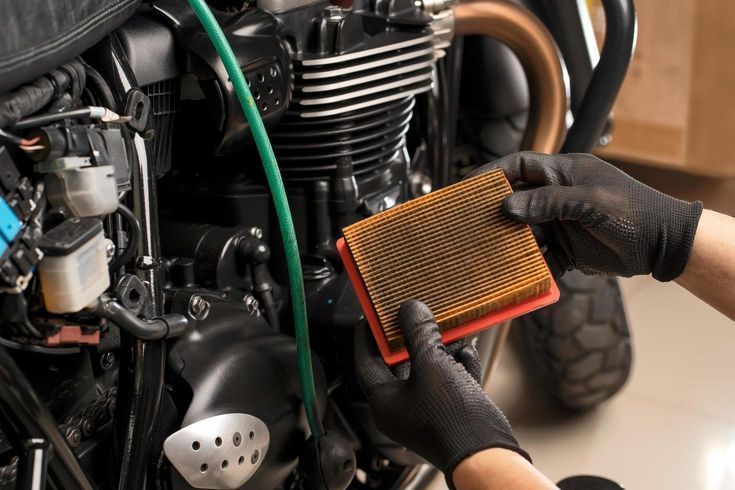 Hands in black gloves holding a motorcycle air filter near an engine.